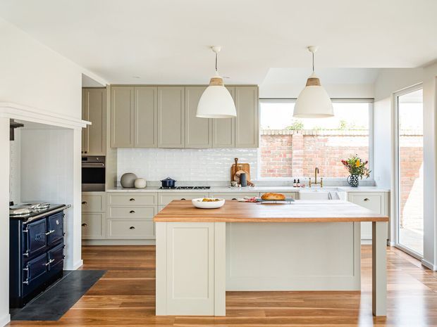 Bright, spacious kitchen with a large central island, shaker-style cabinetry, white tiled splashback, timber benchtop, timber flooring, and pendant lighting. A dark freestanding range cooker sits to the left, while large glazed doors and windows bring in natural light and reveal a brick boundary wall outside.