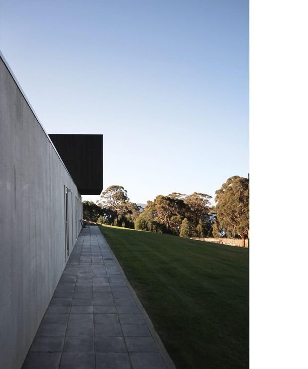 A minimalist modern building exterior viewed along a long paved side walkway. The facade is a smooth grey concrete wall with narrow doors or windows, capped by a projecting dark rectangular volume. A broad lawn and mature trees extend beside the building under a clear sky.