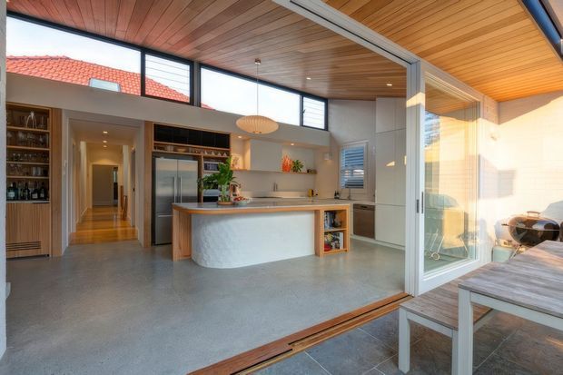 Light-filled contemporary kitchen opening onto an outdoor dining area through large glazed sliding doors. The space features a curved island with timber benchtop and textured light-coloured base, integrated cabinetry, stainless-steel appliances, open shelving, polished concrete flooring, and a timber-lined ceiling with clerestory glazing. A hallway and built-in bar or pantry shelving are visible beyond the kitchen.