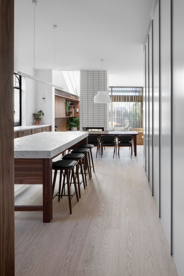 A bright, contemporary open-plan kitchen and dining space viewed along a long circulation area. The kitchen features a large marble-look island with dark timber cabinetry and multiple black bar stools, while a timber-lined display niche and planting add warmth. A white tiled feature wall, sculptural pendant light, pale timber flooring, full-height white cabinetry, and expansive glazed doors create an airy, refined interior.