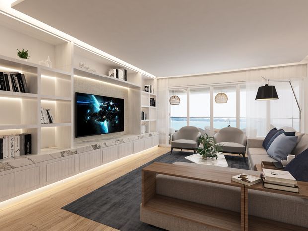 Bright contemporary living room with a full wall of illuminated built-in shelving and cabinetry surrounding a television, pale timber flooring, a dark area rug, and a neutral upholstered sofa and lounge chairs. Large glazing opens to a balcony or terrace with a visible coastal water outlook. Pendant lights, layered LED joinery lighting, and a timber coffee table complete the refined fit-out.