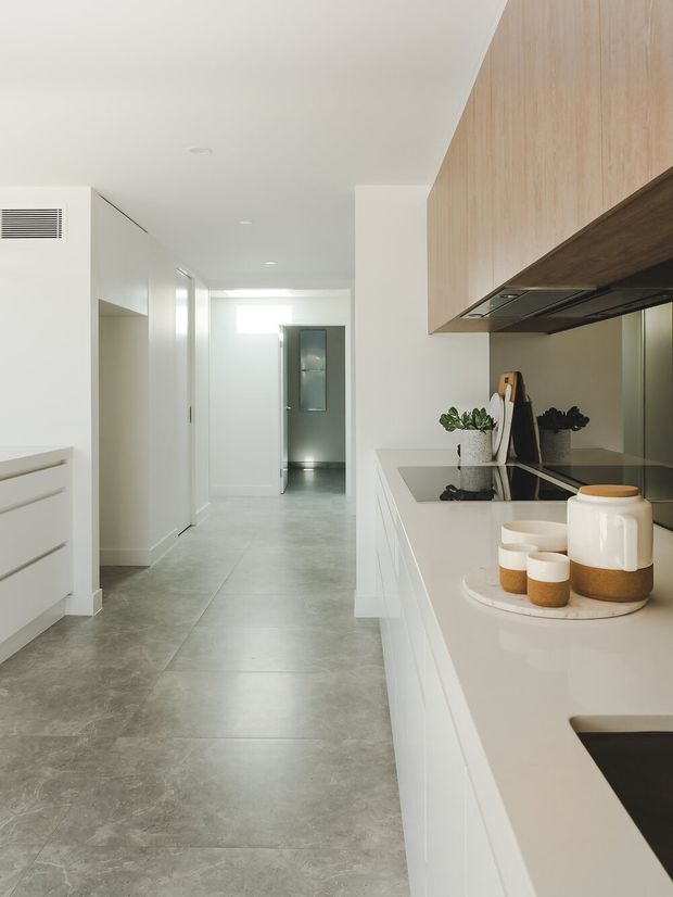 A bright, minimalist contemporary kitchen viewed along a long hallway. White cabinetry and a pale stone-look benchtop frame a black induction cooktop, with light timber overhead cabinetry and integrated rangehood. Grey large-format floor tiles extend through the kitchen and corridor. Small potted plants, a cutting board, and neutral ceramic canisters provide restrained styling.