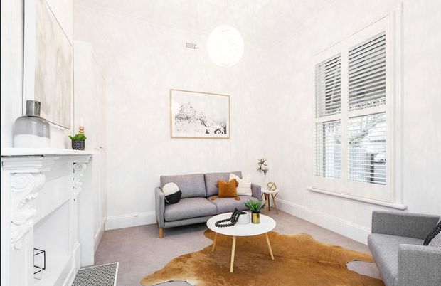 Bright, compact living room with high ceilings, pale textured walls, ornate white fireplace mantel, grey sofa and armchair, round white coffee table, and a large window fitted with white plantation shutters. A cowhide rug, framed artwork, and small decorative plants add warmth and character. The room has a refined, contemporary styling within a traditional architectural shell.