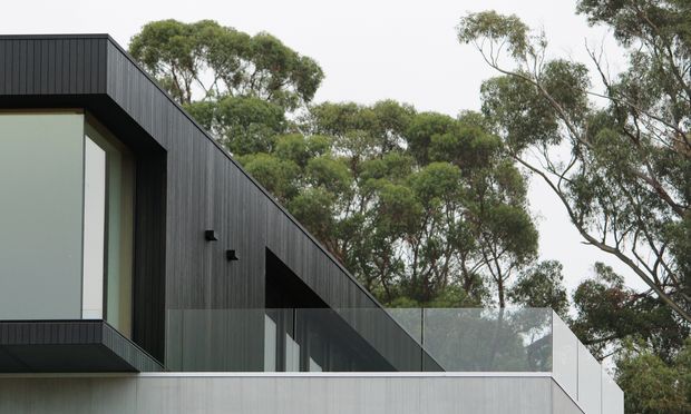 Exterior view of a contemporary, flat-roofed house set among mature eucalyptus trees. The building features dark vertically ribbed cladding, expansive floor-to-ceiling glazing, projecting box-like volumes, and a transparent glass balustrade along an upper terrace or balcony.