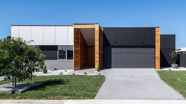 Contemporary single-storey residential facade with a strong geometric composition. The home combines dark charcoal cladding, pale grey upper panels, warm timber-lined entry portal and garage surround, extensive horizontal glazing, and a broad concrete driveway. A simple lawn and young planted tree frame the street-facing exterior under a clear blue sky.