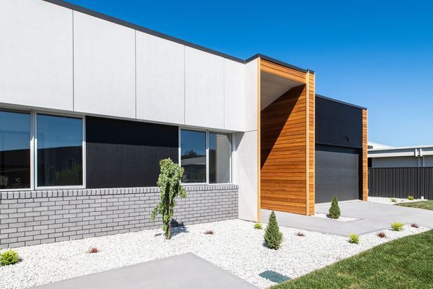 Street-facing contemporary single-storey home facade under a clear blue sky. The exterior combines white panel cladding, dark charcoal sections, a low grey brick base, extensive glazing, and a projecting timber-lined entry recess. A dark garage door sits to the right, while the front garden uses white gravel, sparse clipped planting, concrete paving, and lawn.