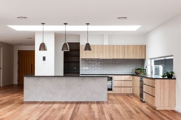 Bright contemporary kitchen with a large concrete-look island, warm timber cabinetry, dark grey brick-pattern splashback, timber flooring, pendant lights, skylight, and an adjoining dining or living area visible beyond the kitchen.
