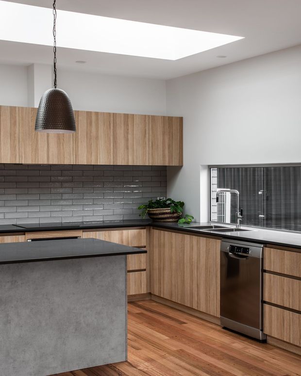 Contemporary kitchen featuring warm timber cabinetry, a dark charcoal tiled splashback, dark benchtops, a concrete-look island front, stainless-steel dishwasher, and timber flooring. A large rectangular skylight provides abundant natural light, while a textured pendant light hangs above the island. A window above the sink adds further daylight.