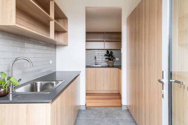 A compact contemporary kitchen viewed toward a secondary kitchen or pantry area. The space features light timber cabinetry and open shelving, a dark stone benchtop, stainless-steel sink and tapware, pale grey subway-tile splashbacks, and grey stone-look floor tiles. A timber-framed glazed door is visible on the right, with natural light creating a bright, clean atmosphere.
