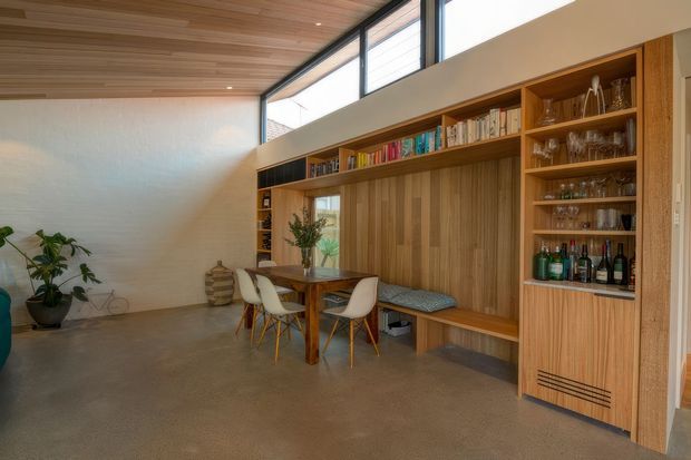 Open-plan dining and lounge area with a built-in timber wall unit incorporating bookshelves, a window seat, display shelving, and a concealed bar or drinks cabinet. A timber dining table with white chairs sits in front of the joinery. The room has a polished concrete floor, timber-lined ceiling, high clerestory glazing, and warm natural materials.