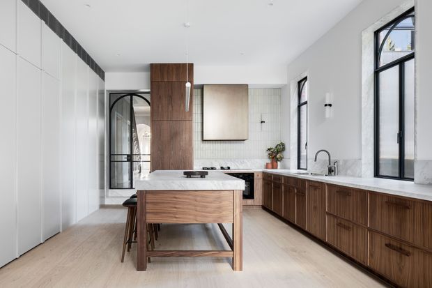 Bright, refined contemporary kitchen with a central timber island, extensive dark timber cabinetry, pale stone benchtops, and a light timber floor. Black-framed arched and rectangular windows and a matching glazed internal door add strong graphic architectural detail, while white walls and a tiled splashback keep the space airy.