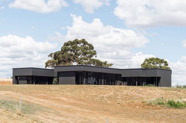 Low, single-storey contemporary building set in an open rural landscape. The dark corrugated-looking facade has a long, horizontally composed form with multiple projecting and recessed wings, flat or very low-pitched rooflines, and extensive dark-framed glazing. Mature trees surround the building, with dry grass and a bright partly cloudy sky in the background.