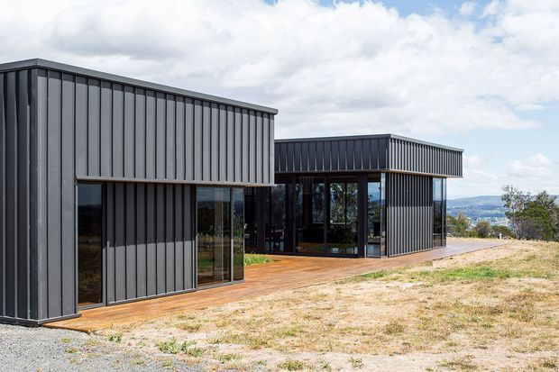 Exterior view of a contemporary single-storey building composed of offset rectangular volumes with dark vertically ribbed metal cladding, extensive floor-to-ceiling glazing, and warm timber decking. The structure sits on a dry, elevated-looking site with distant landscape views and a partly cloudy sky.