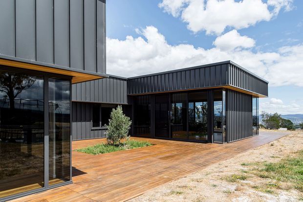 Exterior view of a contemporary, angular multi-volume building with dark vertical metal cladding, extensive floor-to-ceiling glazing, and warm timber decking forming a broad terrace. A recessed glazed entrance sits beneath a projecting flat roof, with a small landscaped planting bed and distant mountainous terrain visible beyond the site.