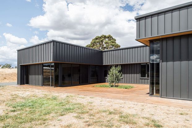 Exterior view of a contemporary single-storey home composed of dark vertical-panel volumes arranged around a sheltered courtyard. Floor-to-ceiling glazing opens onto a broad timber deck, with warm timber soffits and a small landscaped planting bed. The building sits in a dry, open landscape beneath a partly cloudy sky.
