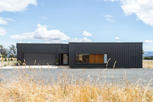 A single-storey contemporary residence with a long, low rectangular form and dark vertically ribbed metal cladding. The front facade includes an integrated dark garage door, a recessed entry, and a wide window with warm timber screening or cladding. The house sits in a sparse, dry landscape with gravel and golden grasses under a bright blue sky; distant water or hills are faintly visible to the right.
