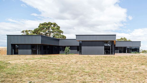 Wide exterior photograph of a low, single-storey contemporary building set in an open grassed site. The building has a strongly horizontal composition with dark grey vertically ribbed metal cladding, large areas of glazing, projecting flat-roofed wings, and simple rectangular volumes. Mature trees and a bright sky form the background.