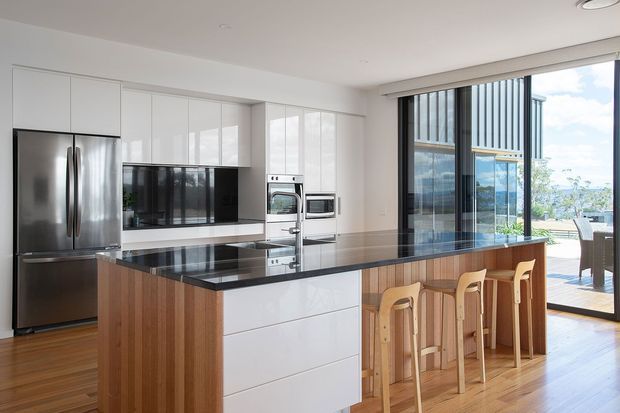 Bright contemporary kitchen with a large central island featuring a polished black stone benchtop, white handleless cabinetry, integrated stainless-steel appliances, and warm timber island panelling. A full-height glazed opening connects to an outdoor terrace and provides views toward a waterfront or coastal setting. Timber flooring, clean lines, and abundant daylight create an airy, refined space.