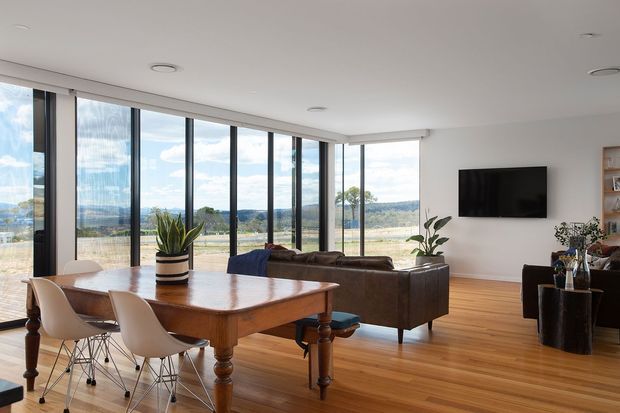 Bright open-plan living and dining area with a large timber dining table, contemporary white dining chairs, leather sofa, wall-mounted television, timber flooring, and expansive floor-to-ceiling glazing overlooking a rural landscape. The room has a clean contemporary fit-out with abundant natural light.