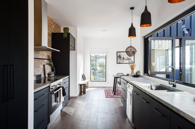 A bright, elongated contemporary kitchen with dark matte cabinetry on both sides, a large pale stone island with an integrated sink, stainless-steel appliances, timber flooring, and a warm exposed brick feature wall. Black pendant lights hang above the island, while large glazed doors or windows bring in abundant daylight and show an adjacent exterior structure. A small dining area sits at the far end.