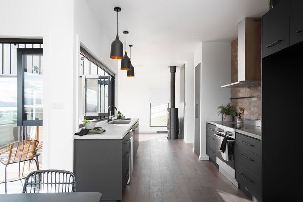 A bright, contemporary galley-style kitchen with a central island, dark charcoal cabinetry, pale stone benchtops, exposed brick splashback, and timber-look flooring. Black pendant lights hang above the island, while large glazed openings bring in natural light and connect to an adjacent outdoor dining area. A freestanding wood-burning stove is visible at the far end of the room.