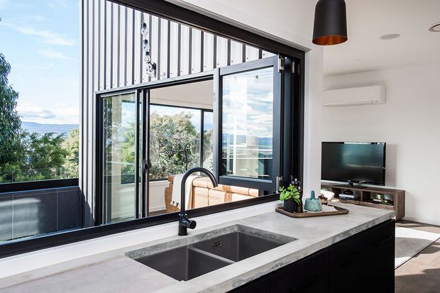 Bright contemporary kitchen interior with a large concrete-look island, integrated double-bowl sink and black cabinetry. Full-height black-framed sliding glazing opens toward a balcony or terrace and reflects a connected outdoor seating area. The room also includes a television and low timber media console, white walls, an air-conditioning unit, and a black pendant light. A leafy landscape and distant water or hills are visible beyond the glazing.