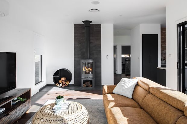 Bright contemporary living room with a tan sectional sofa, round woven coffee table, television and low media console. A freestanding wood-burning fireplace with a dark vertical flue is set against a dark timber feature wall, with firewood stored below and beside it. White walls and ceiling, dark cabinetry, large windows and a glazed external door create a clean, light-filled interior.