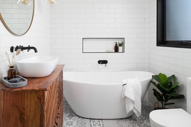 A bright contemporary bathroom featuring a freestanding white bathtub, wall-mounted black tapware, white horizontal subway-tile walls, a timber vanity with slatted detailing and white vessel basin, patterned floor tiles, a recessed display niche, and a wall-mounted television. A large leafy plant and simple accessories add warmth.