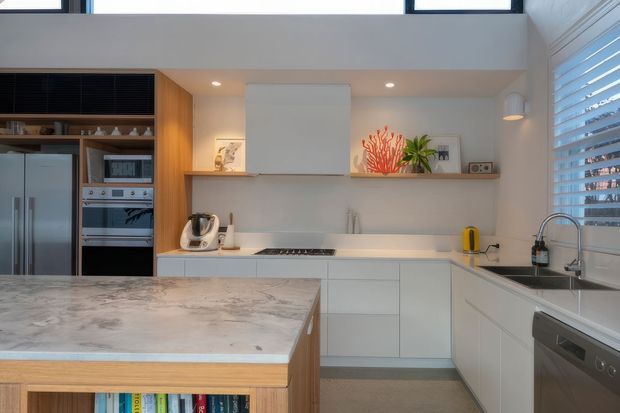 Bright contemporary kitchen with white flat-panel cabinetry, a large pale stone island with timber edging and open shelving, integrated stainless-steel appliances, a white splashback, and a long floating display shelf. Timber cabinetry and warm task lighting add contrast, while plantation shutters and high-level windows bring in natural light.