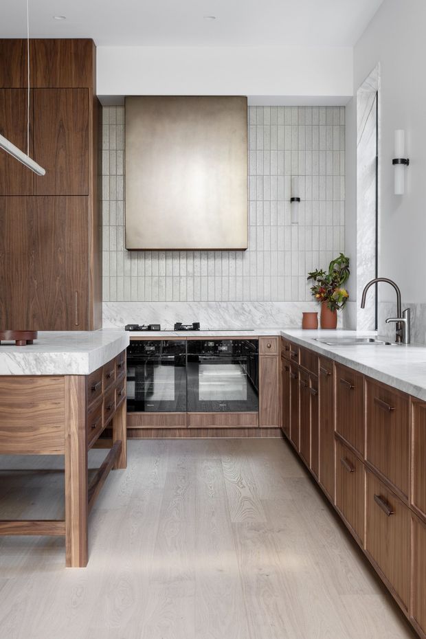 A refined contemporary kitchen with warm walnut cabinetry, pale marble-look stone benchtops, a vertically tiled backsplash, integrated black ovens and cooktop, and a large brushed metal rangehood. A peninsula with open shelving occupies the foreground, while a tall arched faucet and undermount sink sit beneath a bright window. Pale timber flooring and restrained decorative planting complete the light, warm space.