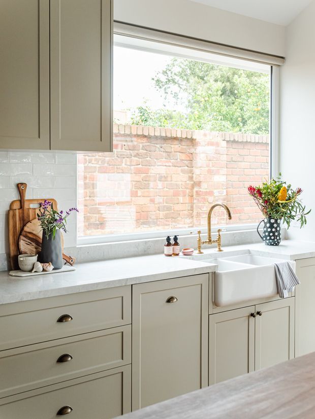 Bright, traditional-style kitchen close-up featuring pale painted shaker cabinetry, a white farmhouse apron-front sink, a light stone benchtop, and a large window overlooking an exposed brick wall. Brass-toned tapware, white subway-tile splashback, timber chopping boards, and a floral arrangement add warm detail.