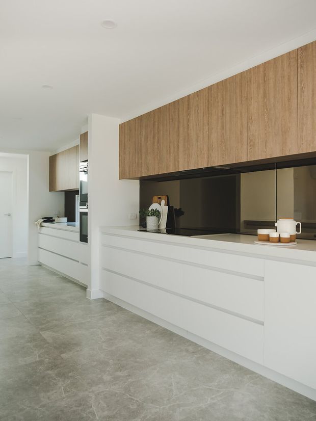 A bright, minimalist contemporary kitchen with a long run of white handleless base cabinetry and warm timber overhead cabinetry. The kitchen features a dark reflective splashback, pale stone-look flooring, integrated appliances, and a clean linear layout extending into the adjoining space.