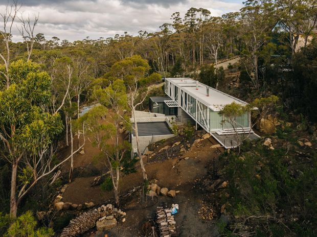 A modern, elongated house set into a heavily wooded and rocky sloping site. The building has a light-coloured flat roof, extensive glazing, slender structural framing, and multiple projecting decks or platforms connected by steps. Native bush surrounds the elevated residence, with exposed rock and rugged terrain clearly visible.