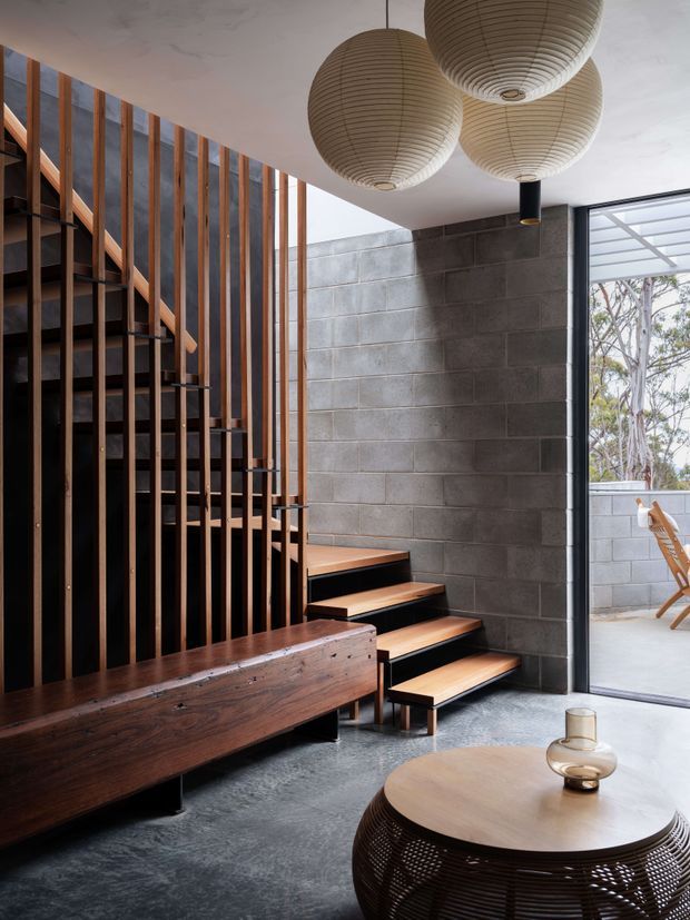 Interior view of a contemporary stairway and living area. A timber-and-black-metal open staircase rises beside a vertical timber slat screen and dark built-in shelving. The wall behind the stairs is finished in exposed grey concrete block, while a large glazed opening leads to an outdoor terrace. Three oversized woven pendant lights hang from the ceiling, and dark timber furniture, a round coffee table, and a low bench occupy the foreground.