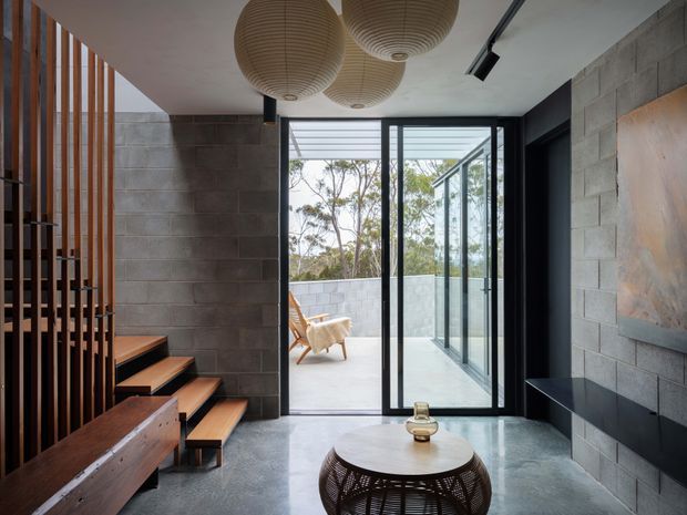 A contemporary industrial-style entry and living area with exposed concrete block walls, polished concrete flooring, black-framed full-height glazing, and timber stair treads behind a vertical timber-screen partition. Woven pendant lights hang above a round coffee table, while the glazing opens to a bright outdoor terrace or balcony.