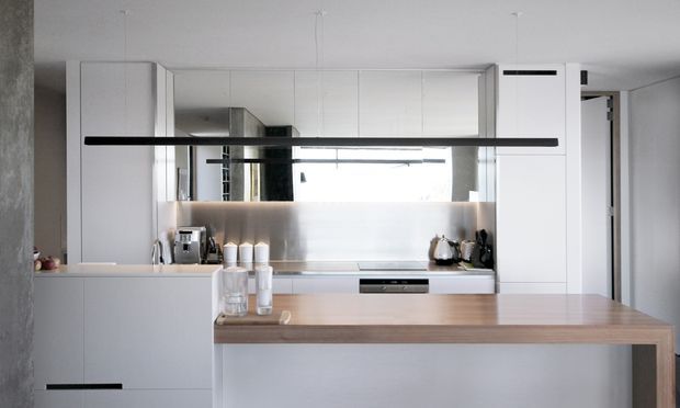 Bright, minimalist contemporary kitchen with white full-height cabinetry, a reflective metallic splashback, pale benchtops, and a timber-topped island. A long linear black pendant light spans the foreground, while integrated appliances and a partially visible dining or circulation area appear beyond the island.