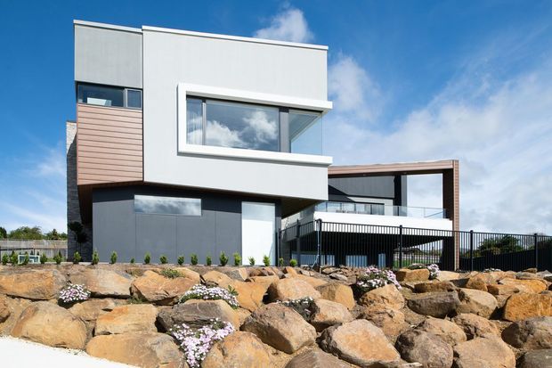 Contemporary multi-level residential home with a strongly geometric facade, flat rooflines, white and charcoal rendered walls, timber-toned vertical cladding, and large glazed openings. The house is viewed from a landscaped rocky frontage with low planting and a fenced upper terrace under a bright blue sky.