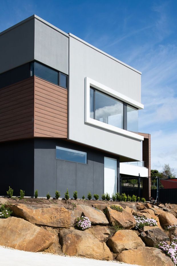 Contemporary multi-level residential house with a strong geometric composition, crisp white and charcoal rendered wall planes, dark aluminium-framed glazing, and warm horizontal timber cladding. The home is built into a rocky sloping site with landscaped stone retaining terraces and small planted areas. A vivid blue sky and clean modern facade create a bright architectural exterior image.