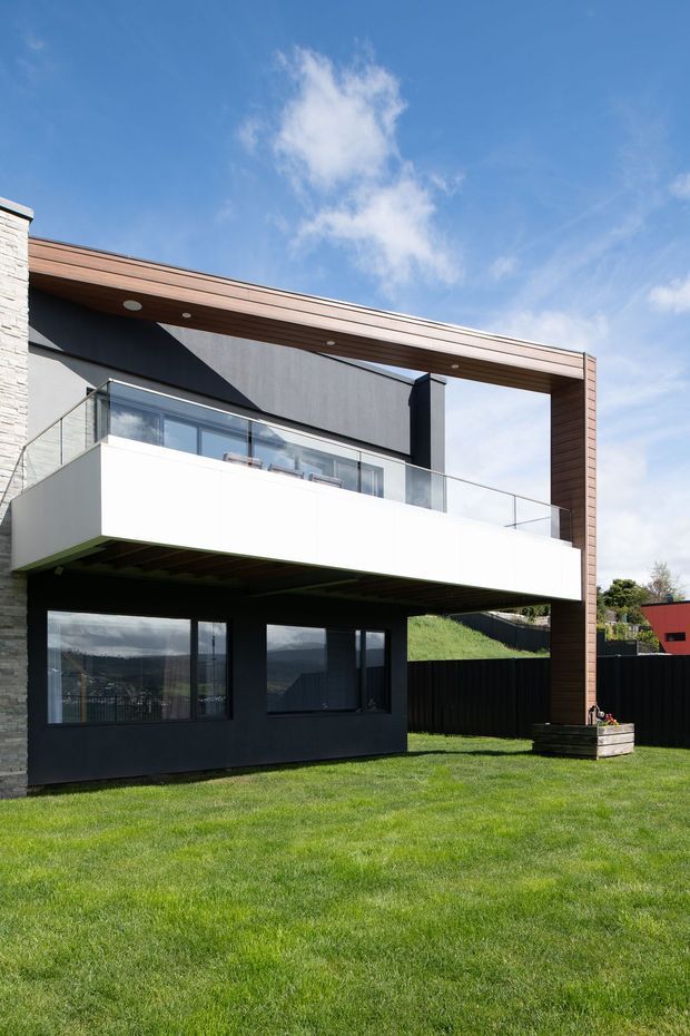 A contemporary multi-level residential house viewed from the rear garden. The facade combines dark cladding, pale stonework, extensive glazing, a projecting white balcony with frameless glass balustrade, and a large timber-lined roof frame or canopy. The lower level is recessed beneath the balcony, while the upper level opens onto the terrace. A broad lawn, dark boundary fencing, and a raised planter sit in the foreground and side yard under a clear blue sky.