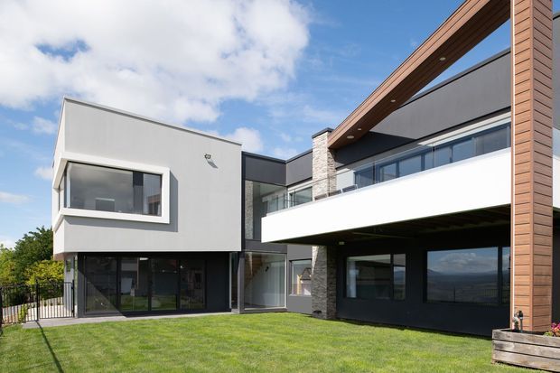 Exterior photograph of a large contemporary two-storey residence arranged around a lawn. The building combines crisp white and dark charcoal rendered volumes, extensive floor-to-ceiling glazing, a projecting upper-level terrace or balcony, a stone-clad vertical feature, and prominent warm timber-lined roof and facade elements. The composition includes strong horizontal and vertical lines, an open covered outdoor area, and a mountain landscape visible beyond the property.