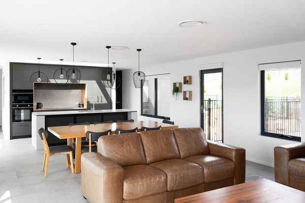 Open-plan contemporary kitchen, dining, and living area with a black-and-white kitchen, central island, timber dining table, pendant lighting, brown leather sofas, and a timber coffee table. Large black-framed windows and a glazed door provide abundant natural light and views to an exterior balcony or deck.
