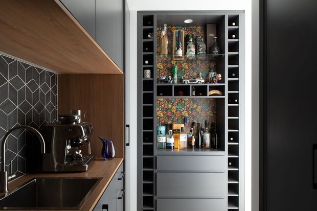 Close-up of a compact contemporary kitchen beverage station beside a sink, featuring dark geometric tiled splashback, warm wood cabinetry and benchtop, a black espresso machine, and a tall built-in drinks cabinet with extensive bottle storage and colourful patterned backing.