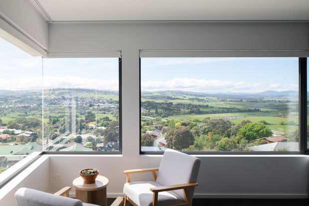 Bright contemporary sitting area with expansive corner windows overlooking a green rural valley and rolling hills. The room features light grey walls, slim dark window frames, pale timber-framed lounge chairs, a small round side table, and a partially visible grey sofa. The glazing and elevated outlook create an airy, calm atmosphere.