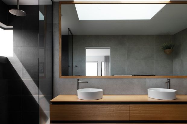 Contemporary double-vanity bathroom with two white vessel basins set on a long timber benchtop and integrated timber vanity cabinetry. A large wood-framed mirror spans the wall above the vanity, reflecting the room and a skylight. The bathroom includes dark charcoal tiled walls and shower enclosure, grey concrete-look wall finishes, black fittings, and a small wall-mounted plant. Strong natural light enters from the skylight, creating a refined, high-contrast interior.
