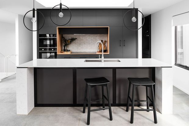 A contemporary kitchen viewed straight on, centred on a large island with white stone surfaces and seating for two. The kitchen features dark matte cabinetry, integrated appliances, a recessed timber-framed display niche with a patterned light splashback, and three black pendant lights. Light grey flooring, white walls, and abundant daylight create a clean, high-contrast interior.