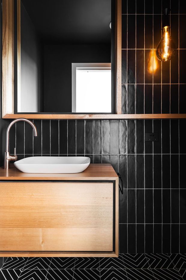 A dramatic, compact bathroom vanity with a white vessel basin set on warm timber cabinetry and benchtop. The walls are clad in glossy black vertically arranged tiles with pale grout, complemented by a black geometric floor tile pattern. A large timber-framed mirror, curved copper-toned tapware, and an exposed warm-glowing pendant add contrast and character.