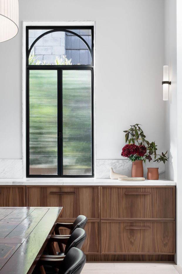 A refined dining area with a built-in walnut-toned sideboard beneath a tall black-framed window. The window combines ribbed translucent glazing with a distinctive arched upper pane, while white marble-look wall cladding and a white wall create a bright backdrop. A dark wood dining table and black upholstered chairs occupy the foreground, with simple wall lighting and decorative vases adding understated detail.