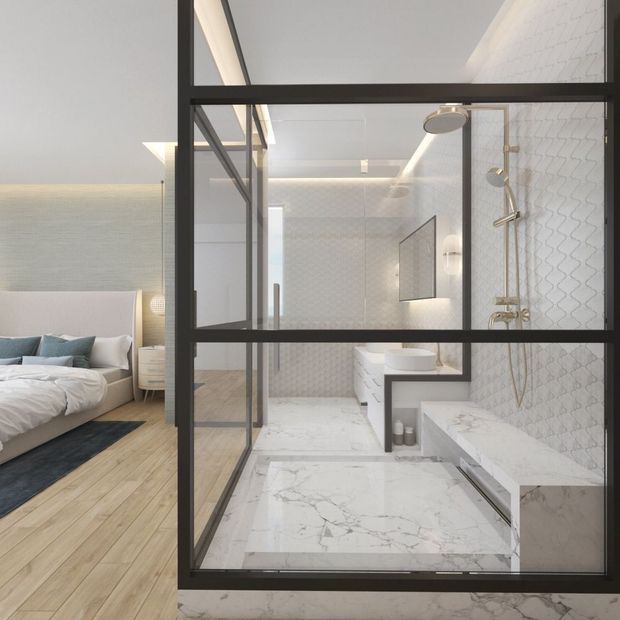 A refined modern bathroom viewed through a black-framed glass partition beside a bedroom. The bathroom features large white marble-look surfaces, a freestanding-style bathtub, wall-mounted vanity, frameless shower fittings, patterned white wall tiles, and brushed metal fixtures. The adjacent bedroom has a low upholstered bed, pale timber flooring, and soft neutral furnishings. The space is bright, clean, and contemporary with strong black-framed glazing defining the bathroom enclosure.