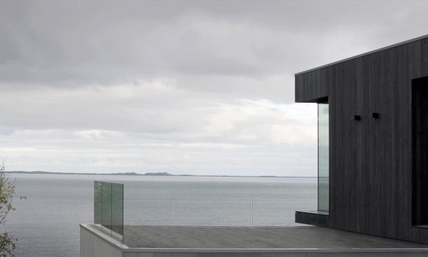 Exterior view of a dark, minimalist contemporary building overlooking a broad body of water. The structure has vertical dark cladding, a strong projecting flat roofline, expansive glazing, and a raised timber-toned terrace enclosed by frameless glass balustrades. The overcast waterfront setting creates a restrained, dramatic atmosphere.