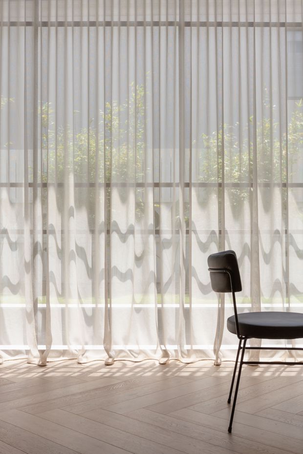 A bright, minimalist interior featuring full-height sheer curtains across a large glazed wall, with soft daylight and leafy greenery visible beyond. The curtains have a subtle vertical striped texture and wavy lower detailing. A simple black upholstered chair sits on a pale timber-look plank floor, creating a calm, airy atmosphere.
